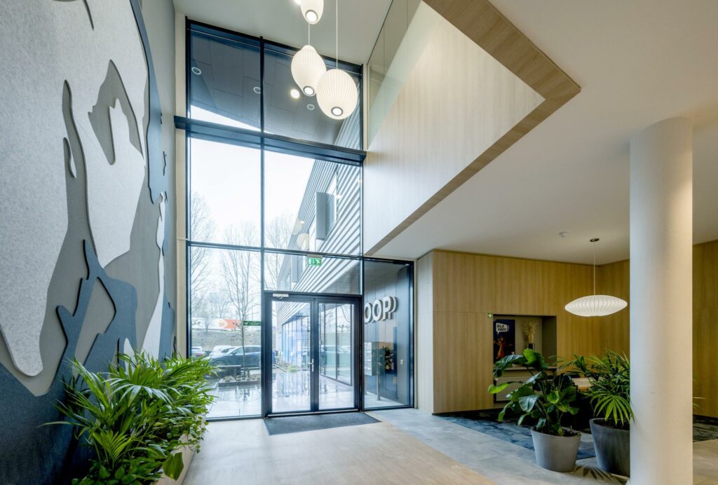Moderne entreehal aan de Joop Geesinkweg met hoge glazen deuren, planten en strakke houten wandafwerking.